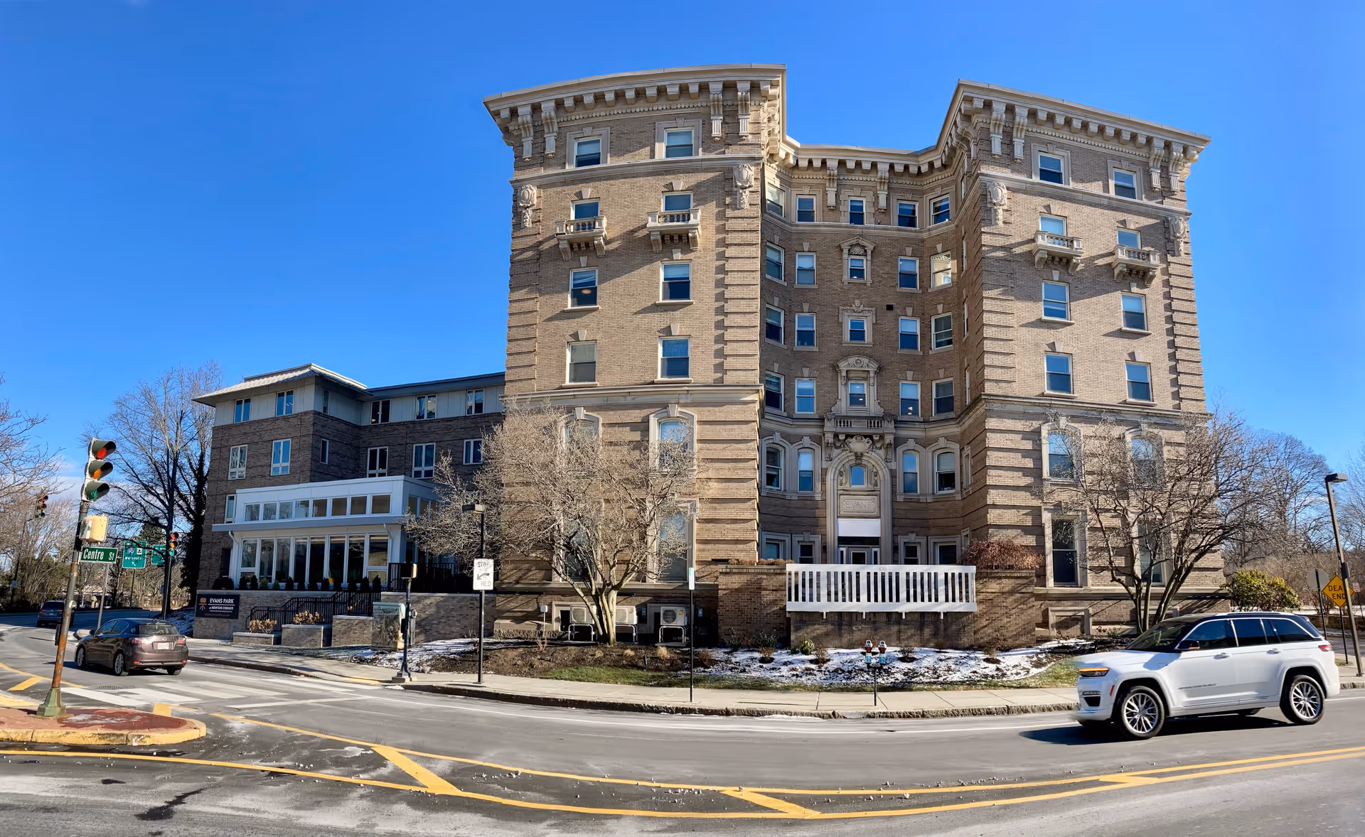 Front exterior of a large multi-story brick senior living building on a street corner under a clear blue sky.