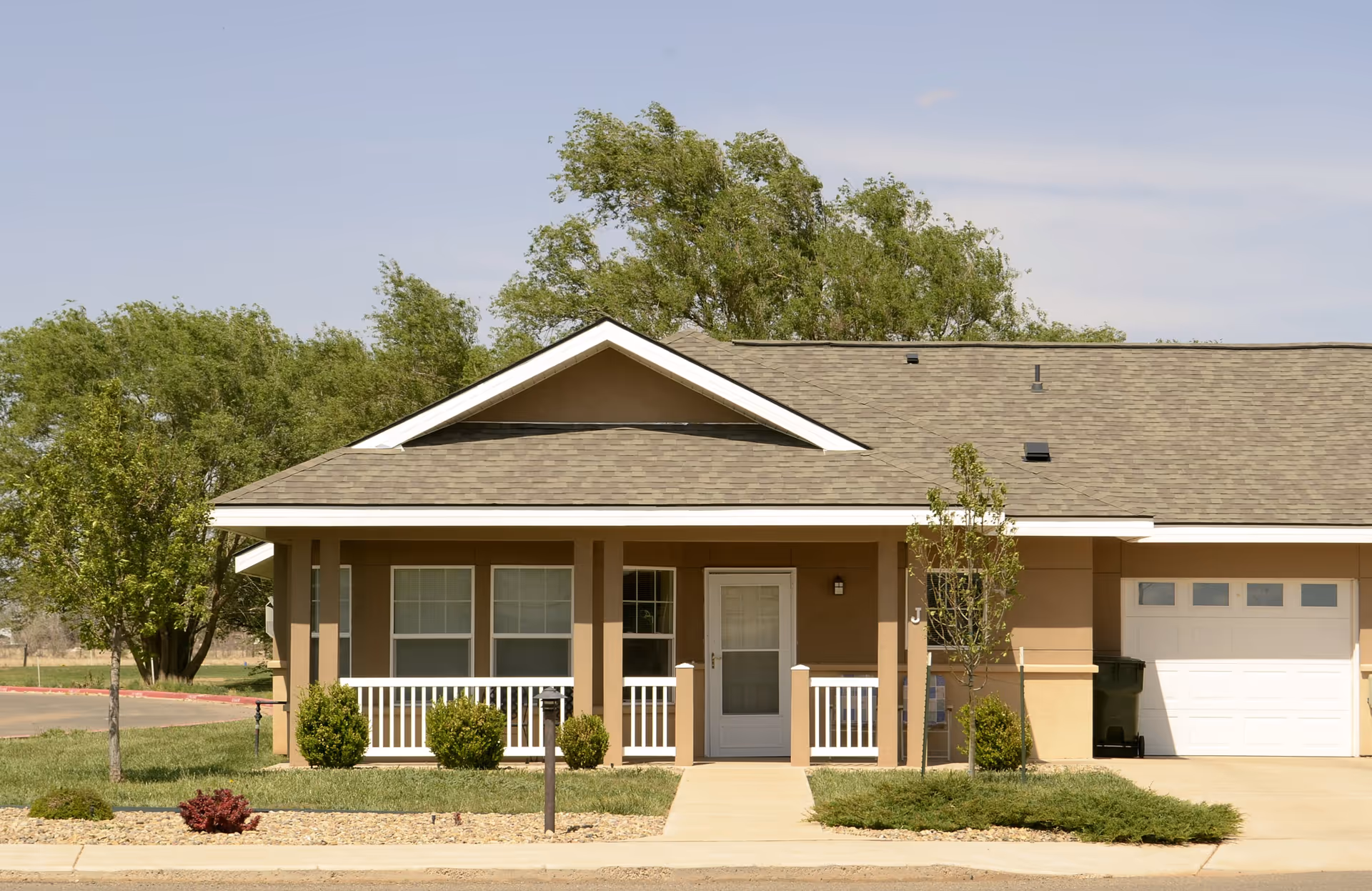 Single-story residential building with a front porch, white railing, and a garage door on the right side. The building is surrounded by small bushes, trees, and a well-maintained lawn under a clear sky.