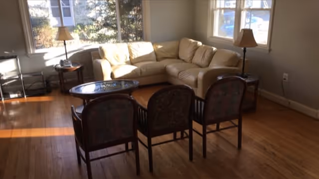 Sunlit living room with a beige sectional sofa, three wooden chairs, and a glass-top coffee table on hardwood floors.