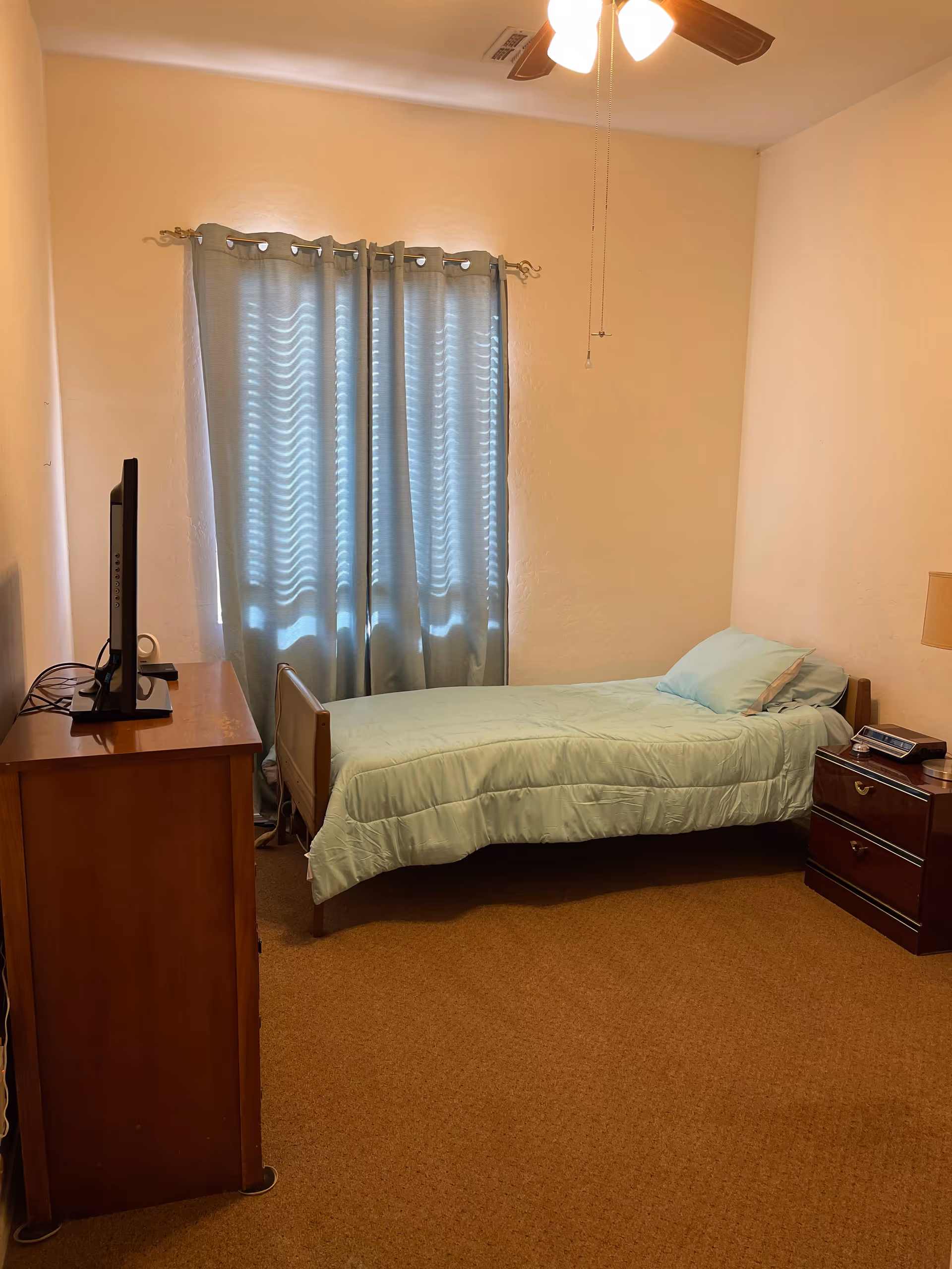A small bedroom with a single bed covered in light blue bedding, a wooden nightstand with a lamp and a clock, a wooden dresser with a flat-screen TV on top, and a window with blue curtains. The room has beige walls and carpeted floor, and a ceiling fan with lights.