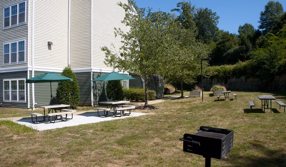 Outdoor area of a senior living facility with picnic tables, some shaded by green umbrellas, a charcoal grill, trees, and a multi-story building with beige and green siding in the background under a clear blue sky.