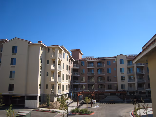 Multi-story Mediterranean-style senior living building with balconies around a central driveway and courtyard under a clear blue sky.