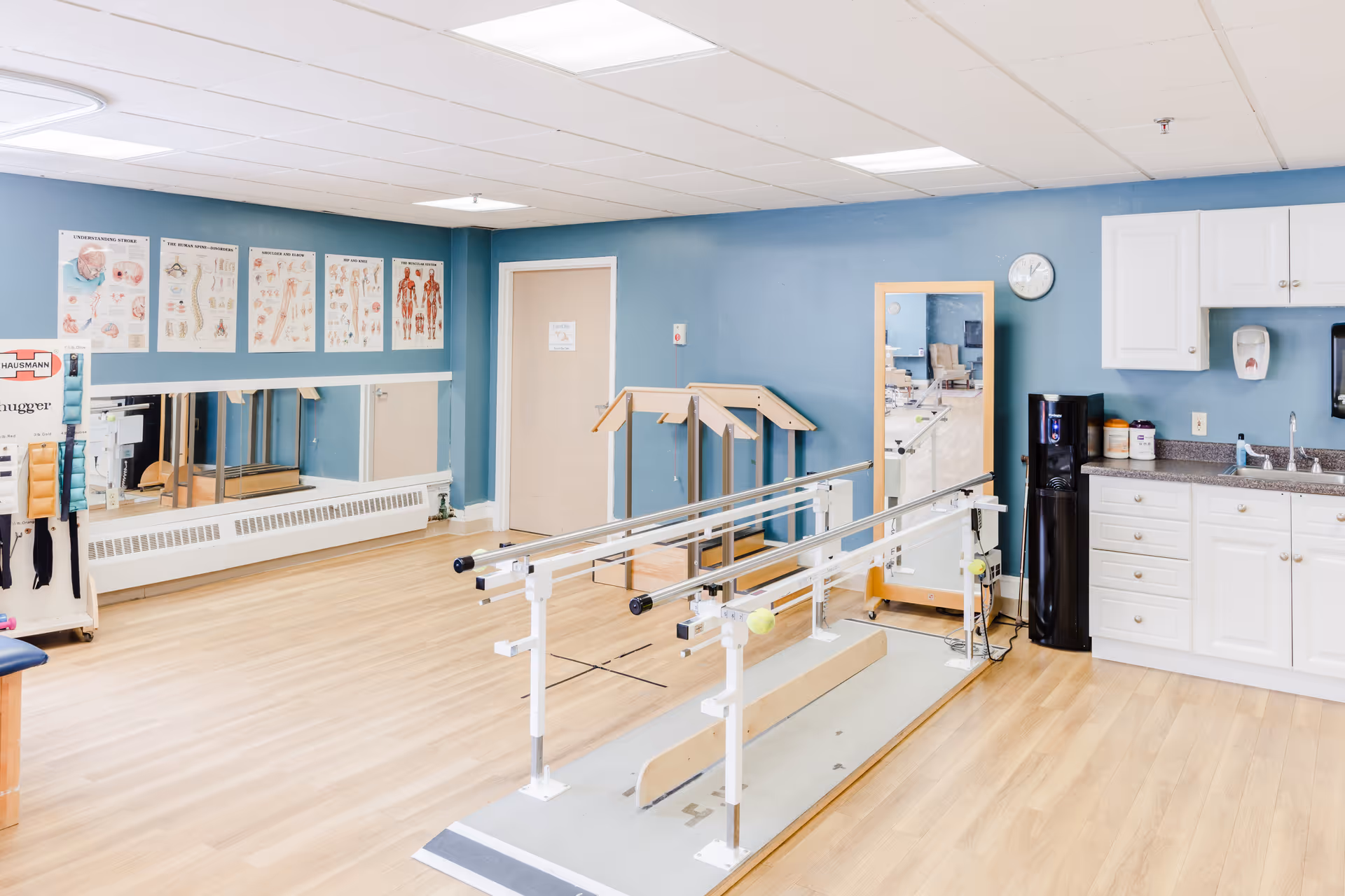 Bright physical therapy/rehab room with parallel bars, mirrors, anatomical charts on the wall, and a small kitchenette area.