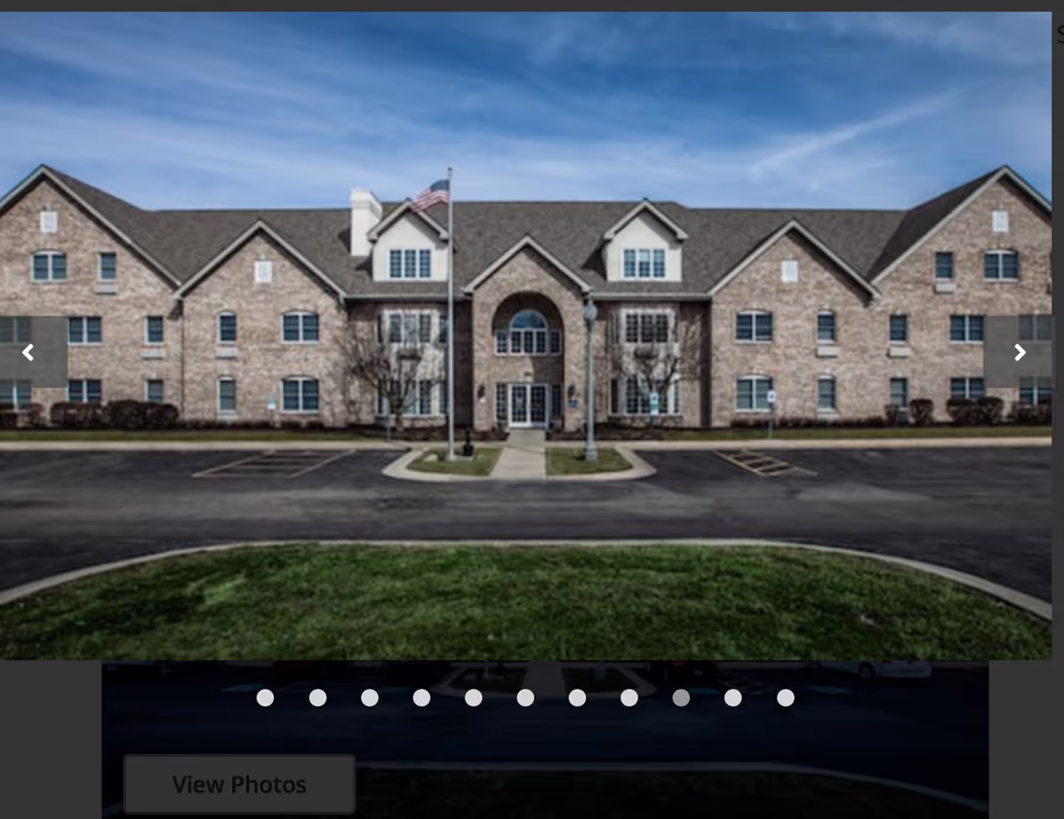 Brick senior living building with central arched entrance, flagpole, and parking lot.