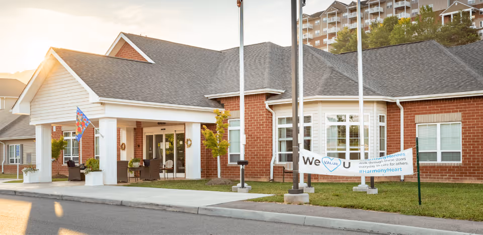 Exterior view of a single-story brick building with a covered entrance, white pillars, and a small porch area with chairs. A colorful flag is displayed near the entrance, and a banner on the lawn reads 'We VALUE U' with additional supportive text. The building is part of a senior living facility with a multi-story building visible in the background.