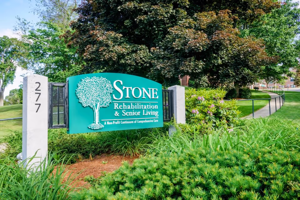 Green entrance sign for Stone Rehabilitation & Senior Living amid landscaped grounds and a paved walkway to the building.