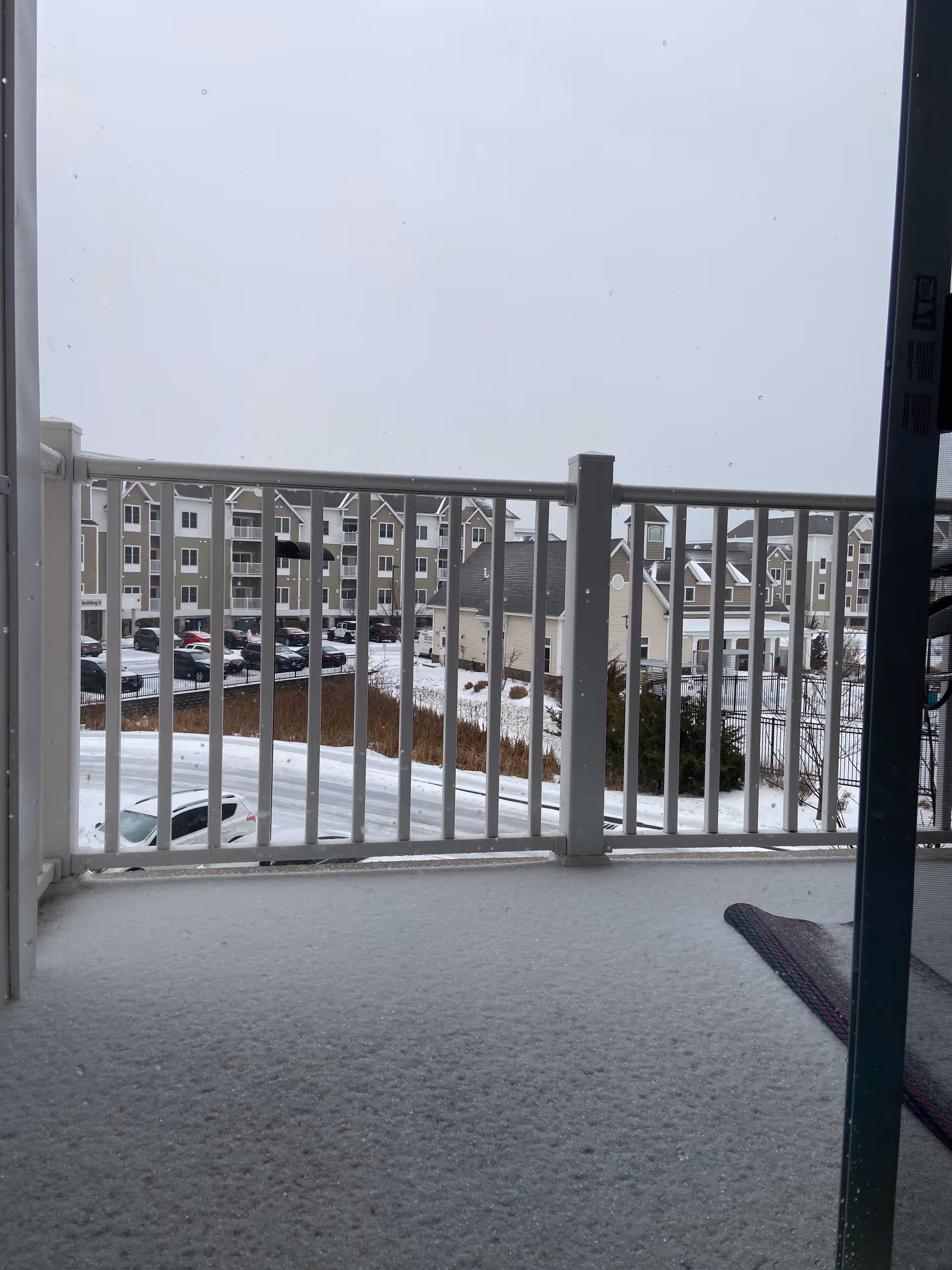 Snowy balcony view overlooking a parking lot and apartment buildings of the facility