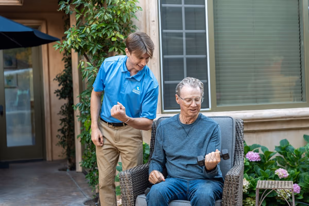 Caregiver in a blue polo chats with an elderly man seated outdoors.