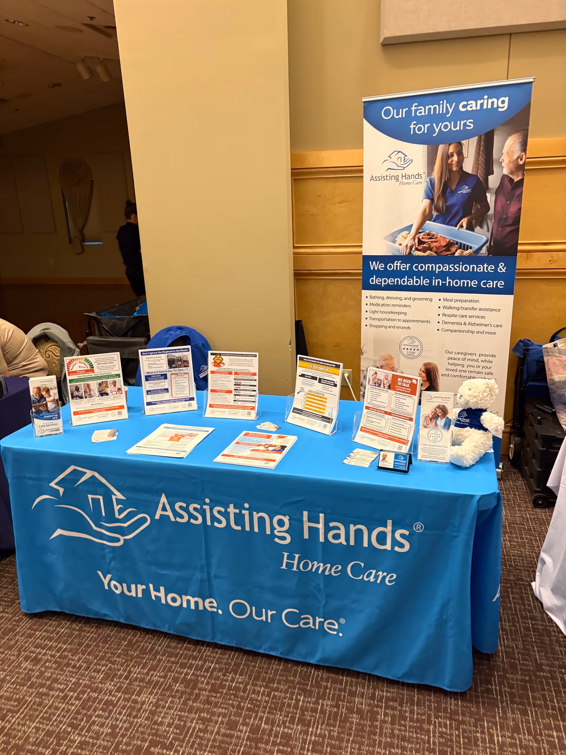 Promotional table with blue cover and banner for Assisting Hands Home Care at an indoor event.
