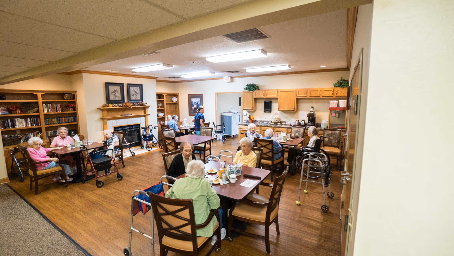 Seniors seated at tables in a communal dining room with a kitchenette, fireplace, bookshelves, and walkers nearby.