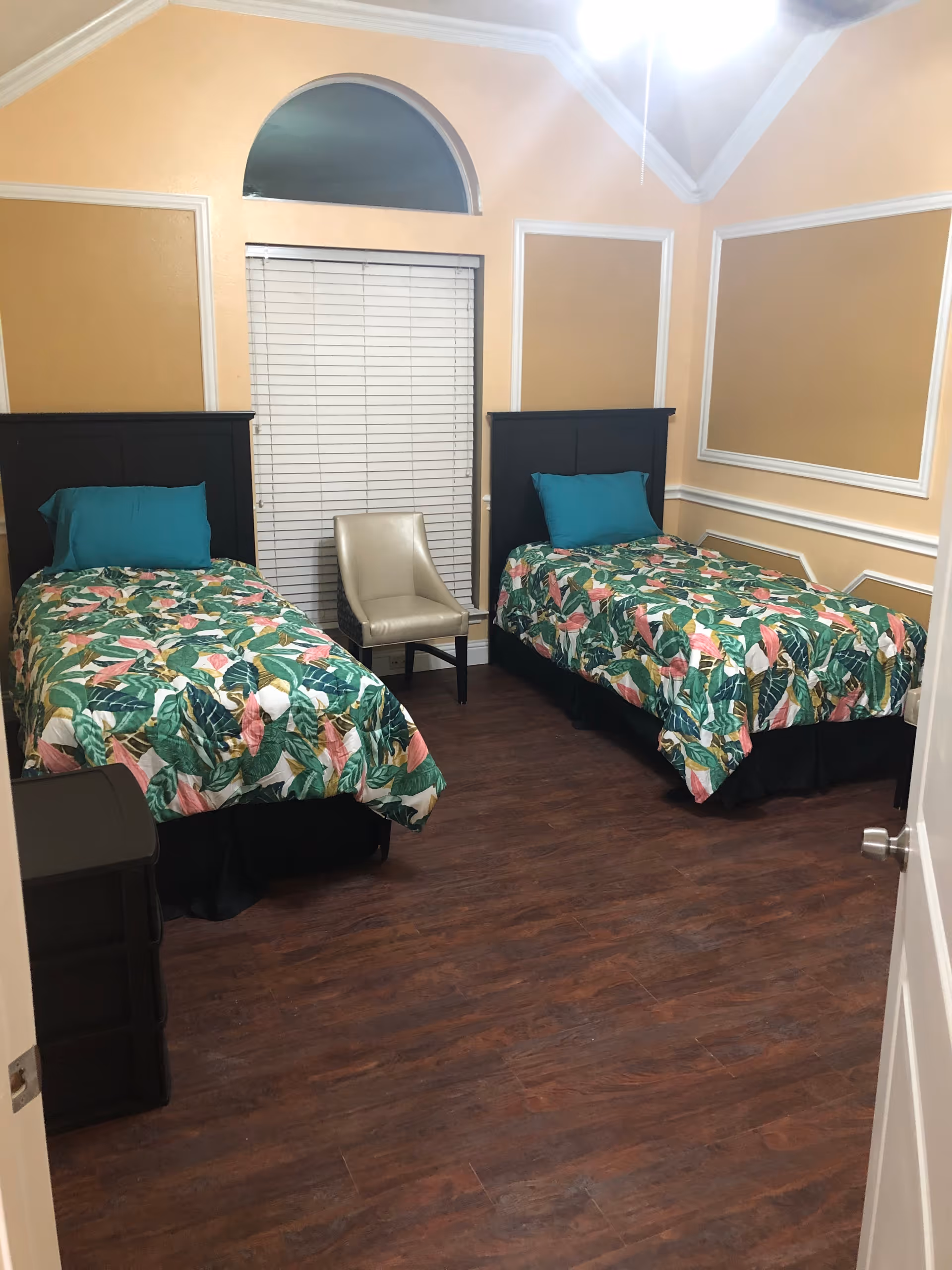 A bedroom with two twin beds, each with a black headboard, teal pillows, and colorful floral bedspreads. Between the beds is a beige chair placed in front of a window with white blinds and an arched window above it. The walls are painted a warm beige color with white trim, and the floor is a dark wood laminate.