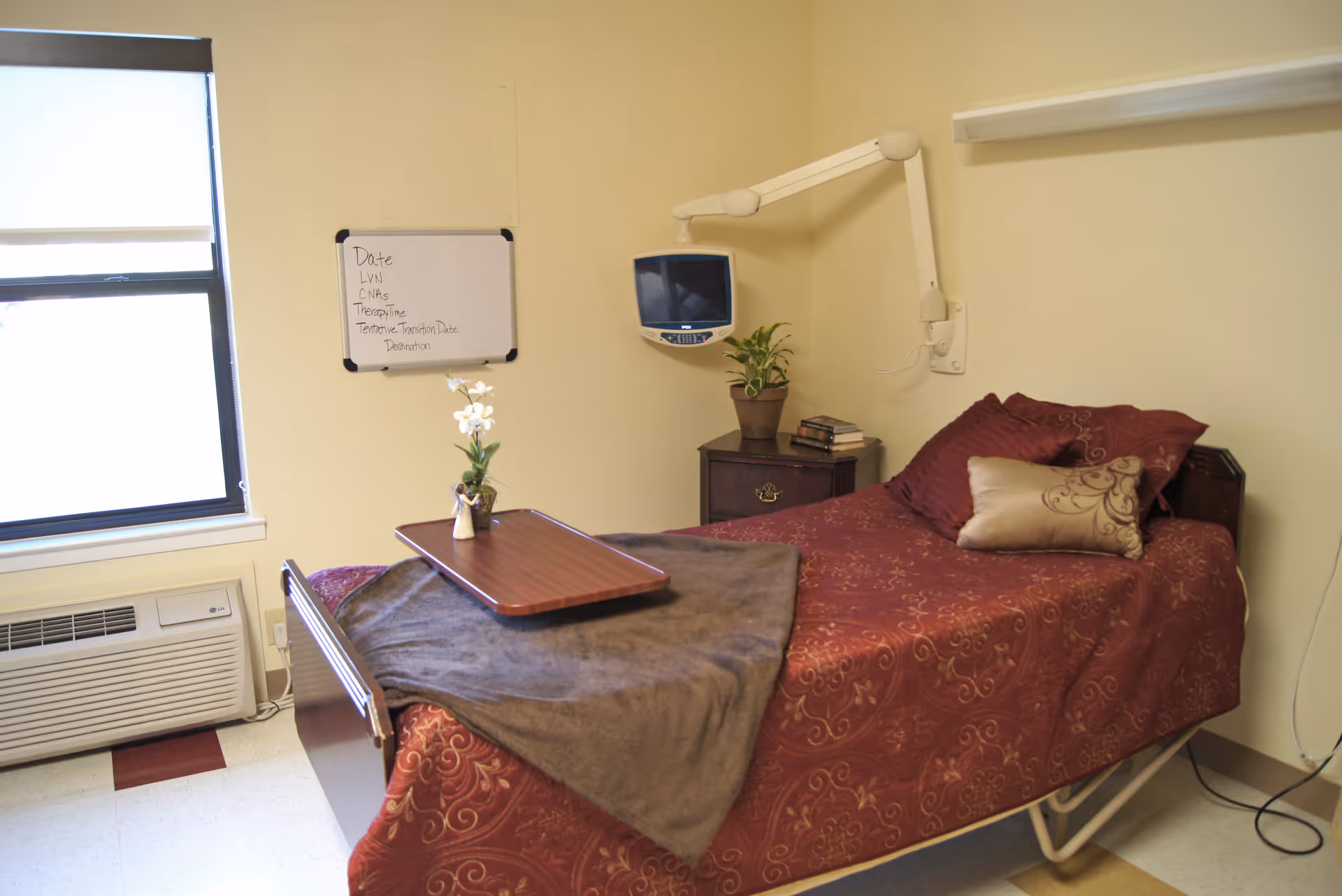 A single bed in a room with a maroon patterned bedspread and decorative pillows. A brown blanket and a wooden tray with a small vase of white flowers are on the bed. Next to the bed is a small nightstand with a potted plant and some books. A wall-mounted TV with an adjustable arm is above the nightstand. A whiteboard with handwritten notes is on the wall near a window with a roller shade. Below the window is an air conditioning unit.