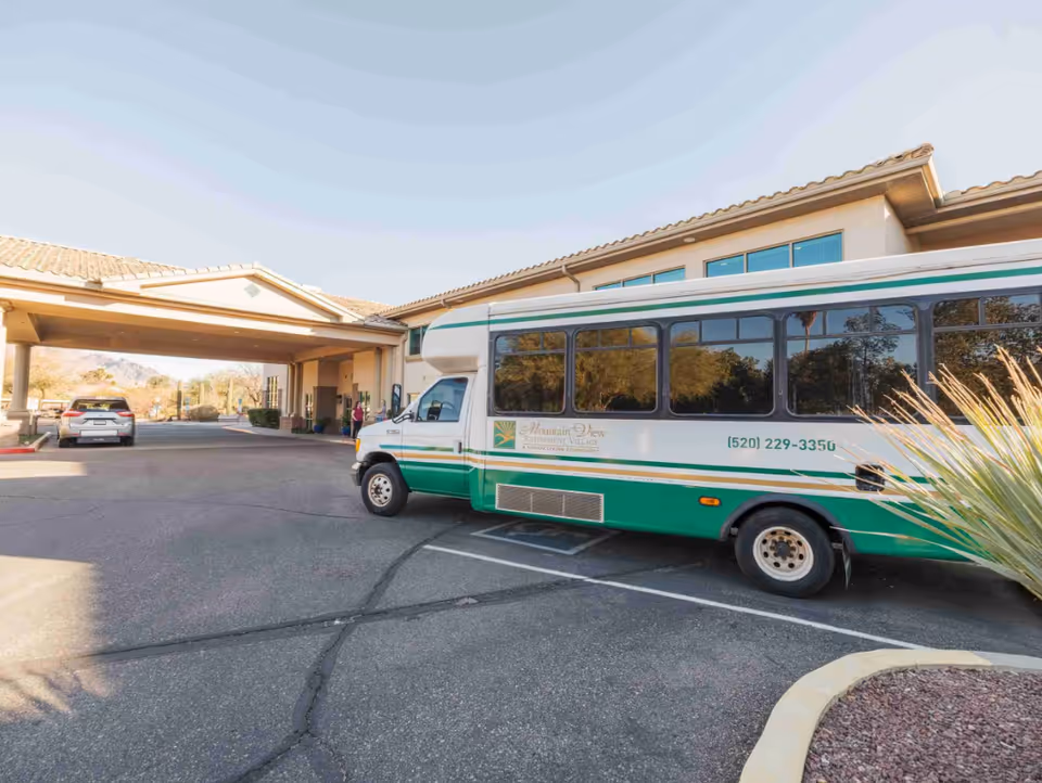 Front entrance and drop-off area of a retirement community with a green-and-white shuttle bus parked near the covered porte-cochère.