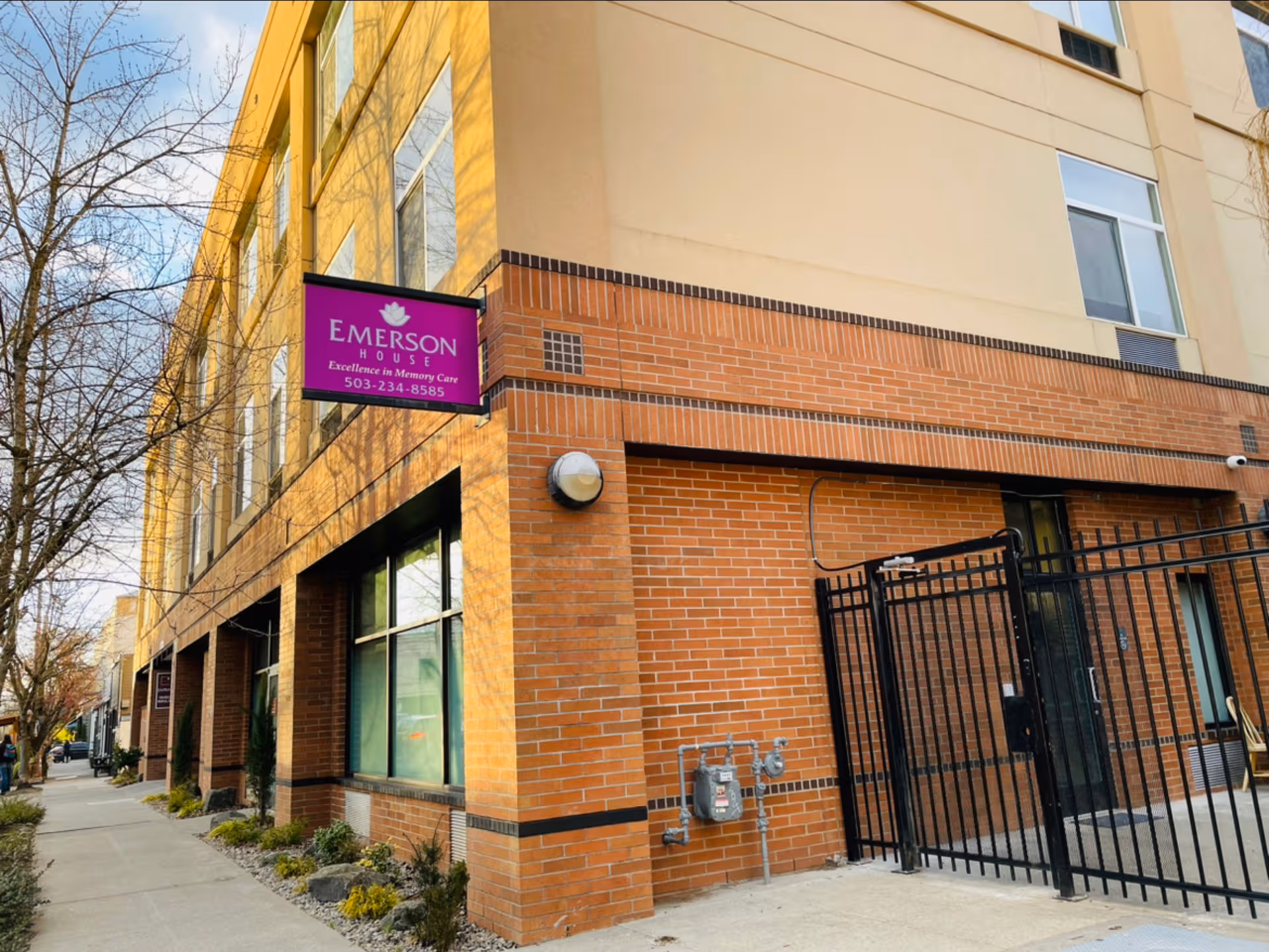 Brick exterior of Emerson House senior living facility with purple sign and sidewalk.