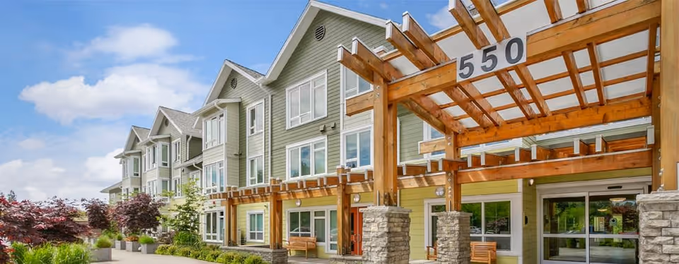 Front exterior of a modern multi-story senior living building with a wooden pergola entrance marked "550", benches, landscaping and large windows.