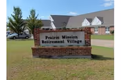 Brick sign for Prairie Mission Retirement Village with a grassy lawn and building in background.