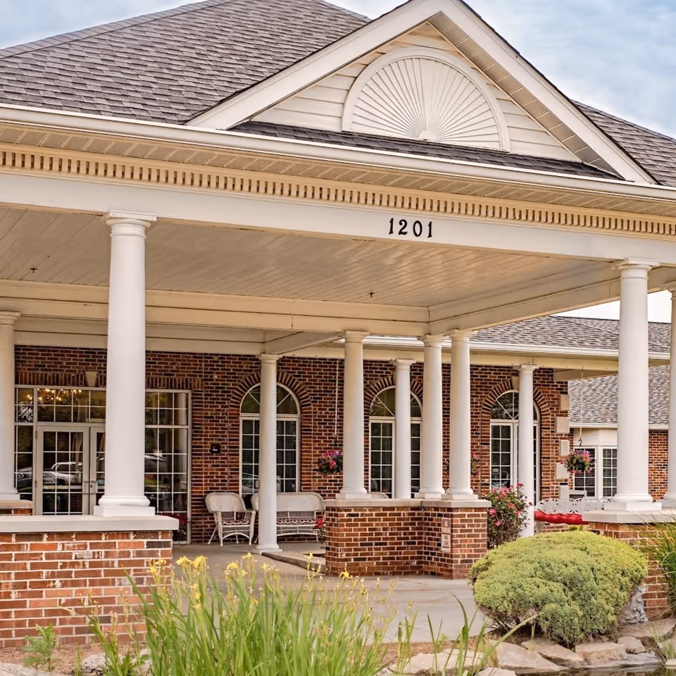 Front exterior view of a senior living facility with white columns supporting a covered entrance. The building has red brick walls, large windows with white frames, and a decorative triangular pediment above the entrance with the number 1201 displayed. There are some bushes and flowers in the landscaped area near the entrance.