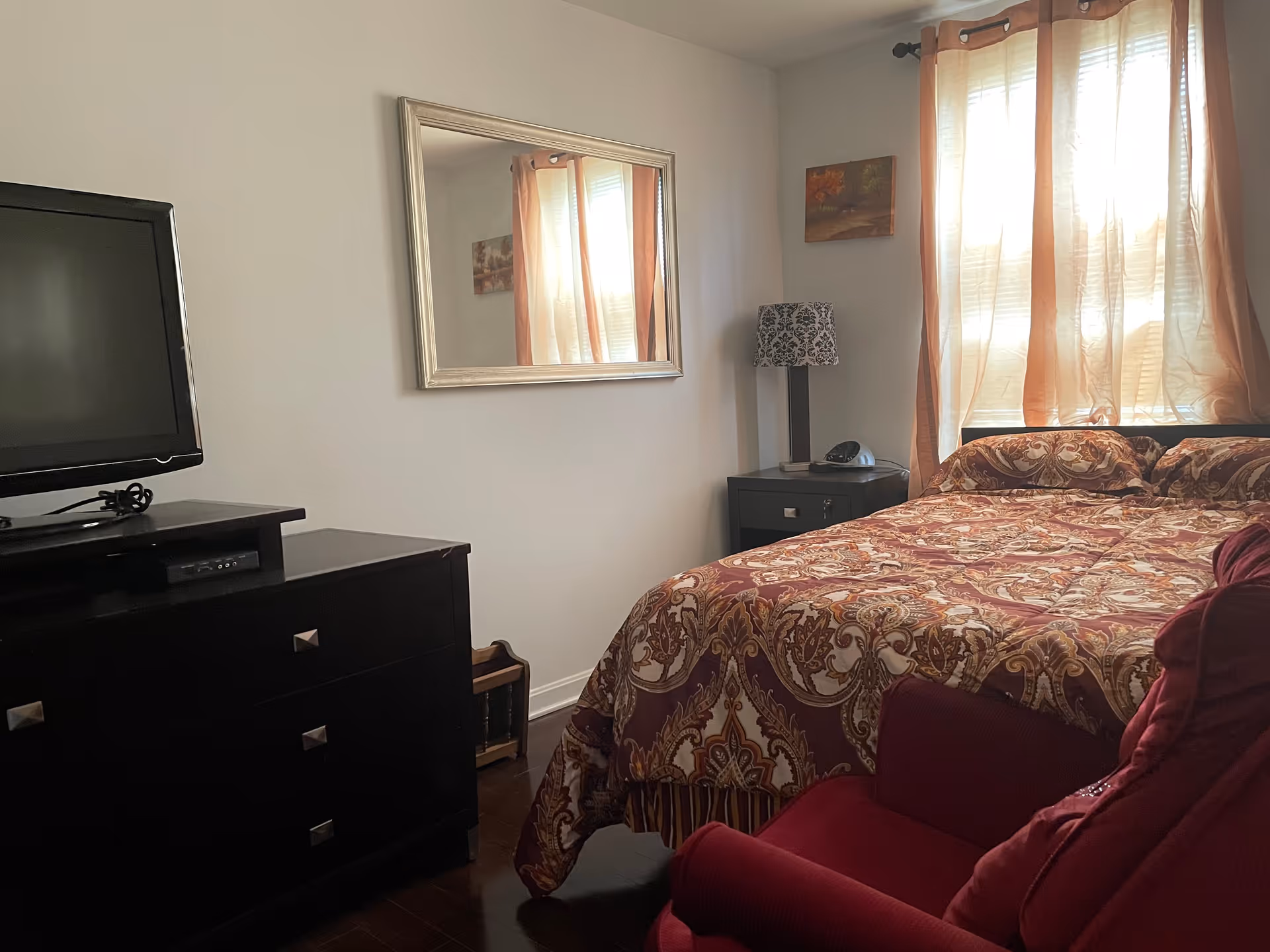 Cozy bedroom featuring a bed, dresser, TV, mirror, curtains, and a red chair.