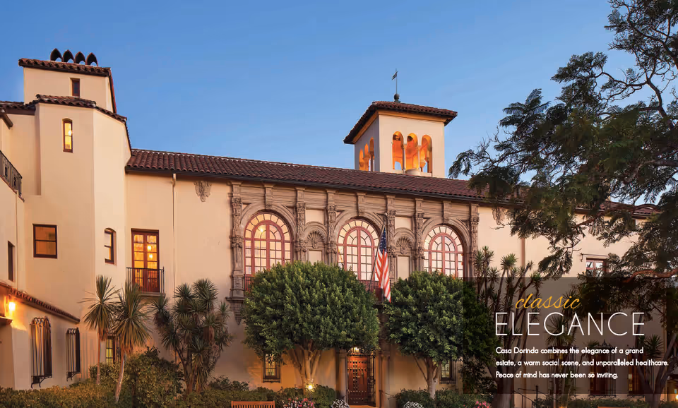 Exterior view of Casa Dorinda, a large building with classic architecture featuring arched windows, a tiled roof, and a bell tower. The building is surrounded by trees and shrubs, with an American flag displayed near the entrance. The sky is clear and blue.