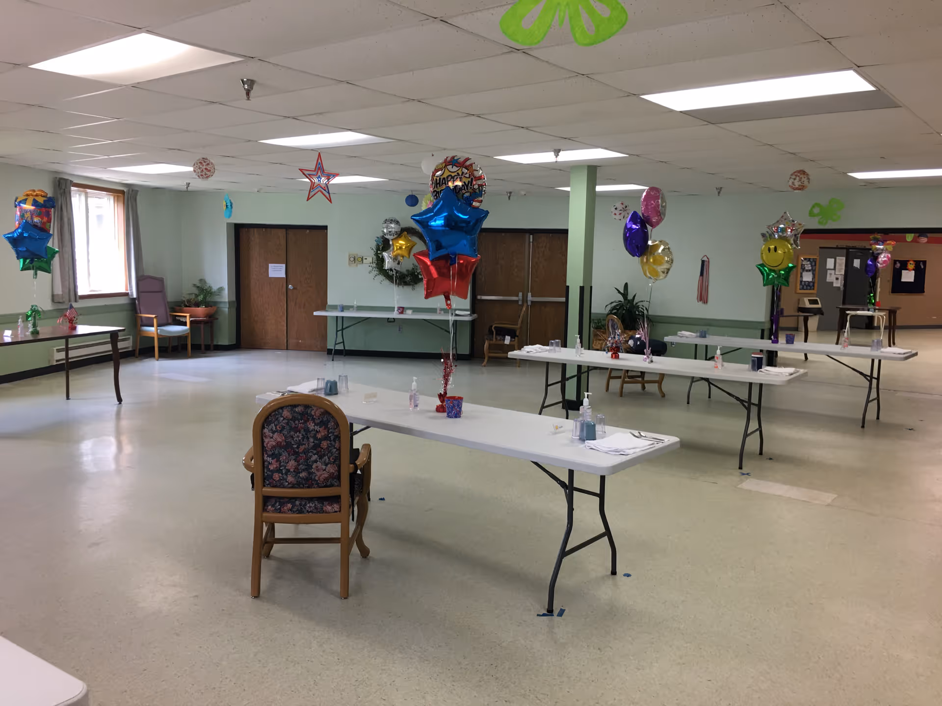 A large, well-lit room with several folding tables spaced apart, each decorated with colorful balloons and set with napkins, utensils, and hand sanitizer bottles. There are a few chairs, potted plants, and festive hanging decorations from the ceiling. The walls are light green with wooden double doors at the back.
