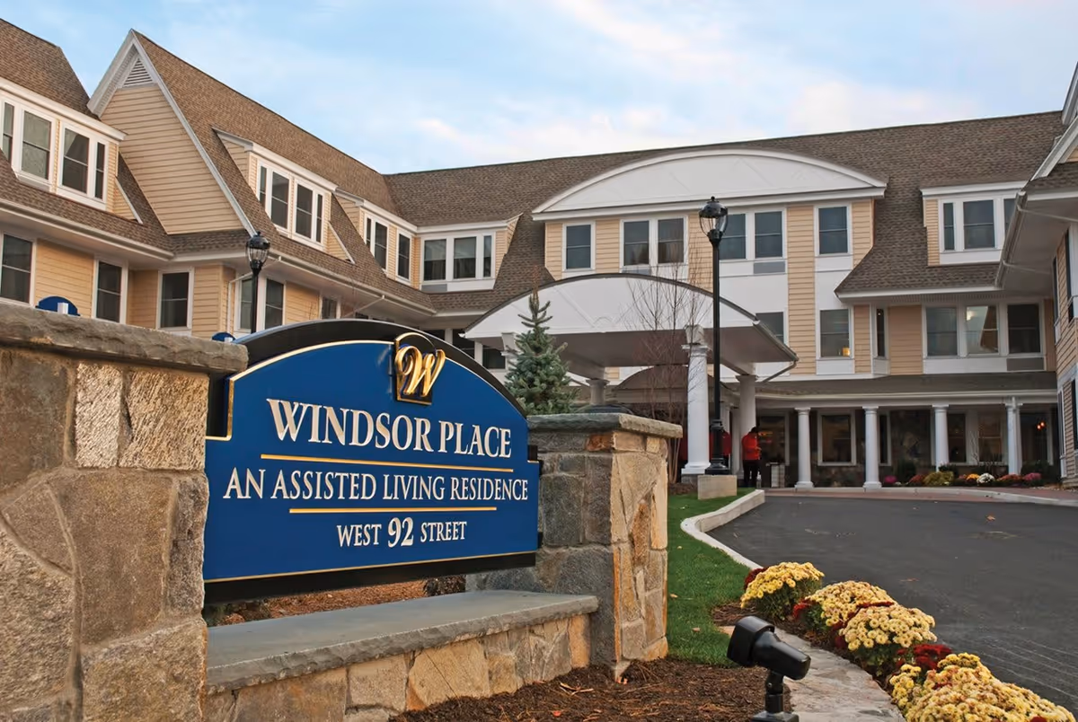 Exterior view of Windsor Place, an assisted living residence, showing the main entrance with a covered drop-off area, a stone sign with the facility name and address, and a landscaped driveway with flowers and shrubs.