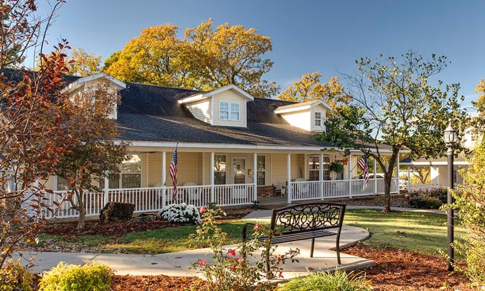 Front exterior of a yellow two-story senior living building with a wraparound porch, American flags, landscaped grounds and a bench in the foreground.