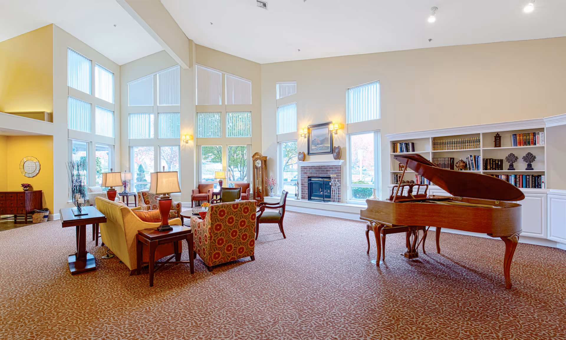 A spacious senior living lounge with a grand piano, seating, bookshelves, and a fireplace.