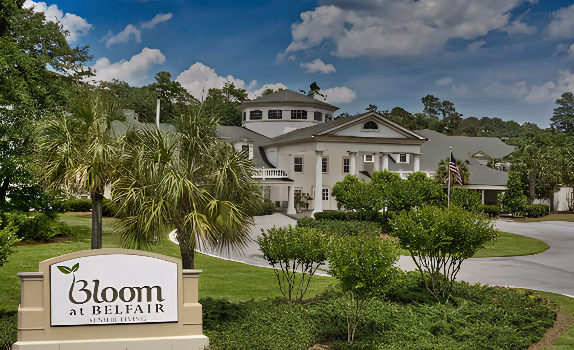 Exterior view of Bloom at Belfair senior living facility showing a large white building with columns, surrounded by green trees and bushes under a partly cloudy blue sky. A sign in the foreground reads 'Bloom at Belfair Senior Living'.