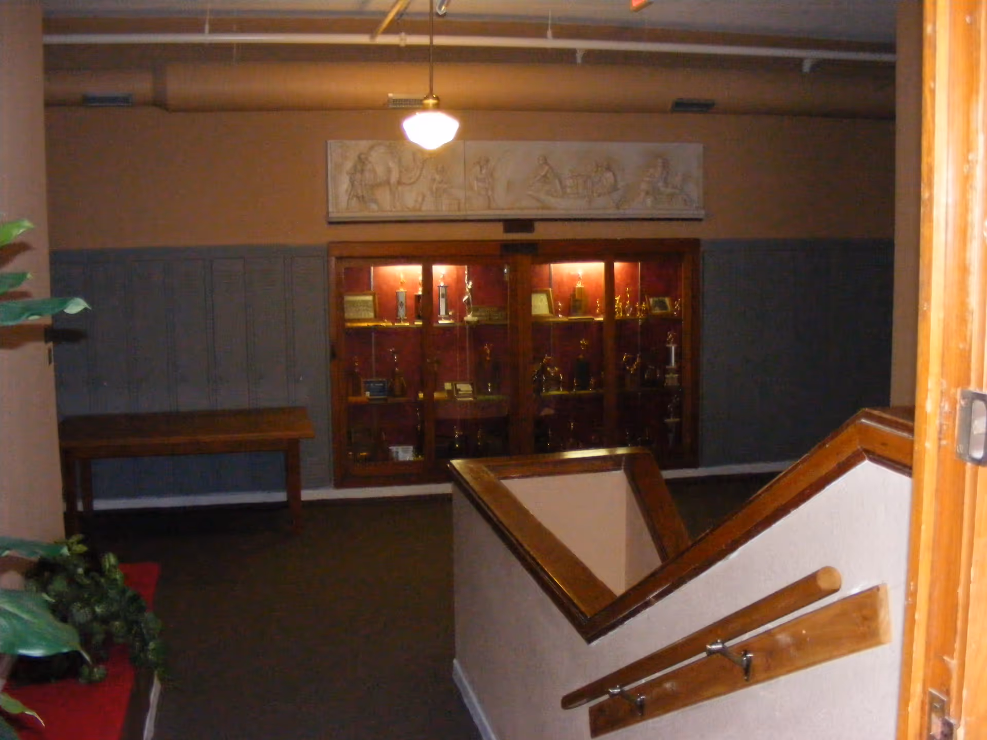 Interior view of a hallway with a staircase railing in the foreground, a wooden table against the wall on the left, and a glass trophy display case illuminated from within on the far wall. Above the display case is a decorative wall panel with carved figures. There are plants partially visible on the left side.