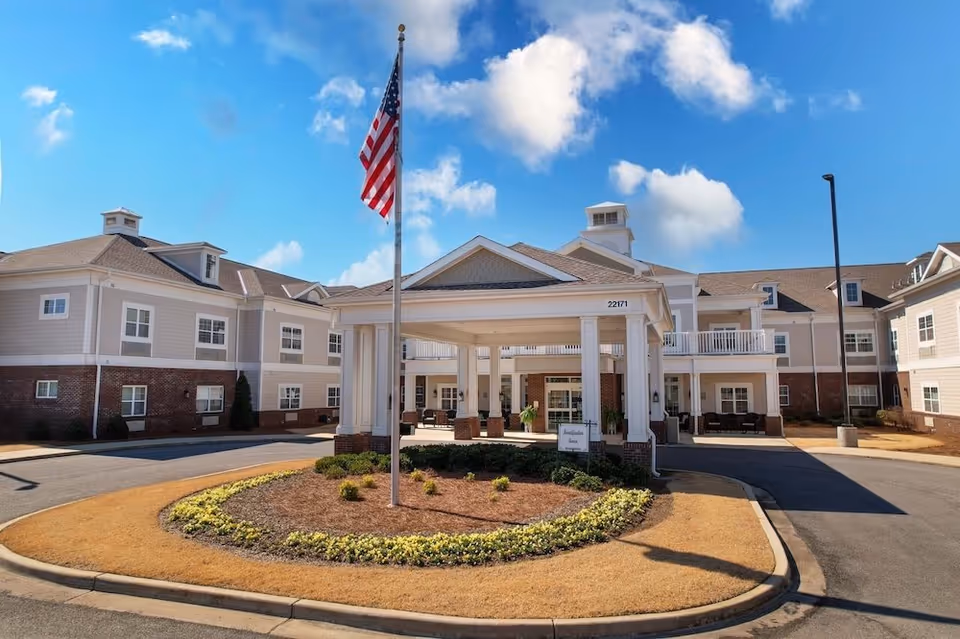 Front entrance of a senior living building with a covered porte-cochere, American flag on a flagpole, circular driveway, and landscaped island.