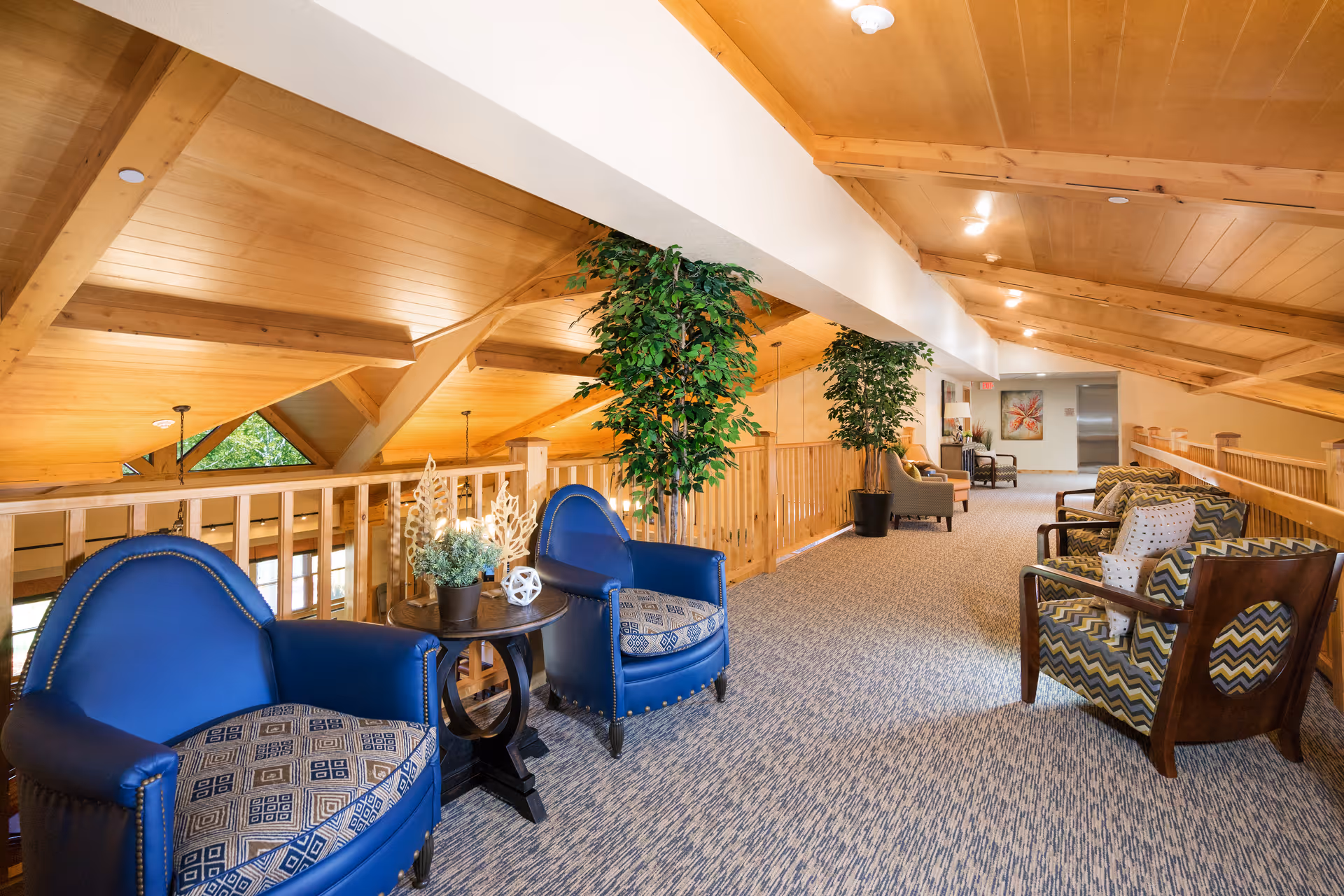 A spacious indoor seating area with wooden vaulted ceilings and railings. The area features several upholstered armchairs in blue and patterned fabric, small tables with decorative plants, and large potted plants along the railing. The space is well-lit with ceiling lights and has a carpeted floor.