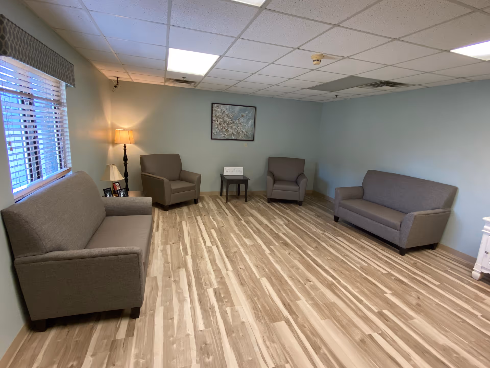 A simple, clean living room with light blue walls and wood-patterned flooring. The room contains two gray sofas and two gray armchairs arranged around a small dark wooden side table. There is a floor lamp and a table lamp in the corner near a window with blinds. A framed floral painting hangs on the wall above the side table.