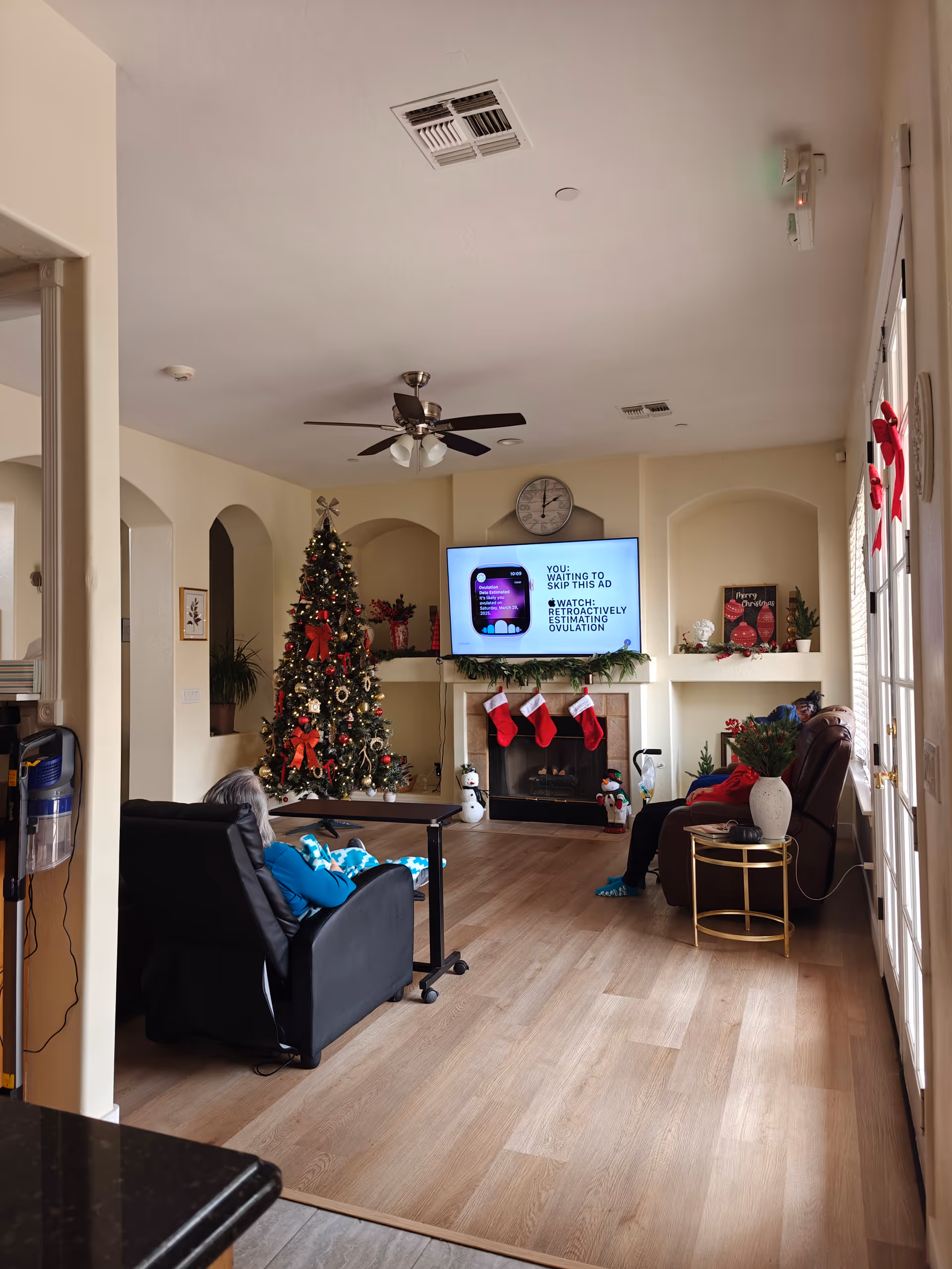 A cozy senior living lounge decorated for Christmas with a tree, stockings, fireplace, and residents.