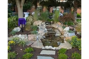 Rock waterfall surrounded by stepping stones and lush garden beds in an outdoor setting.