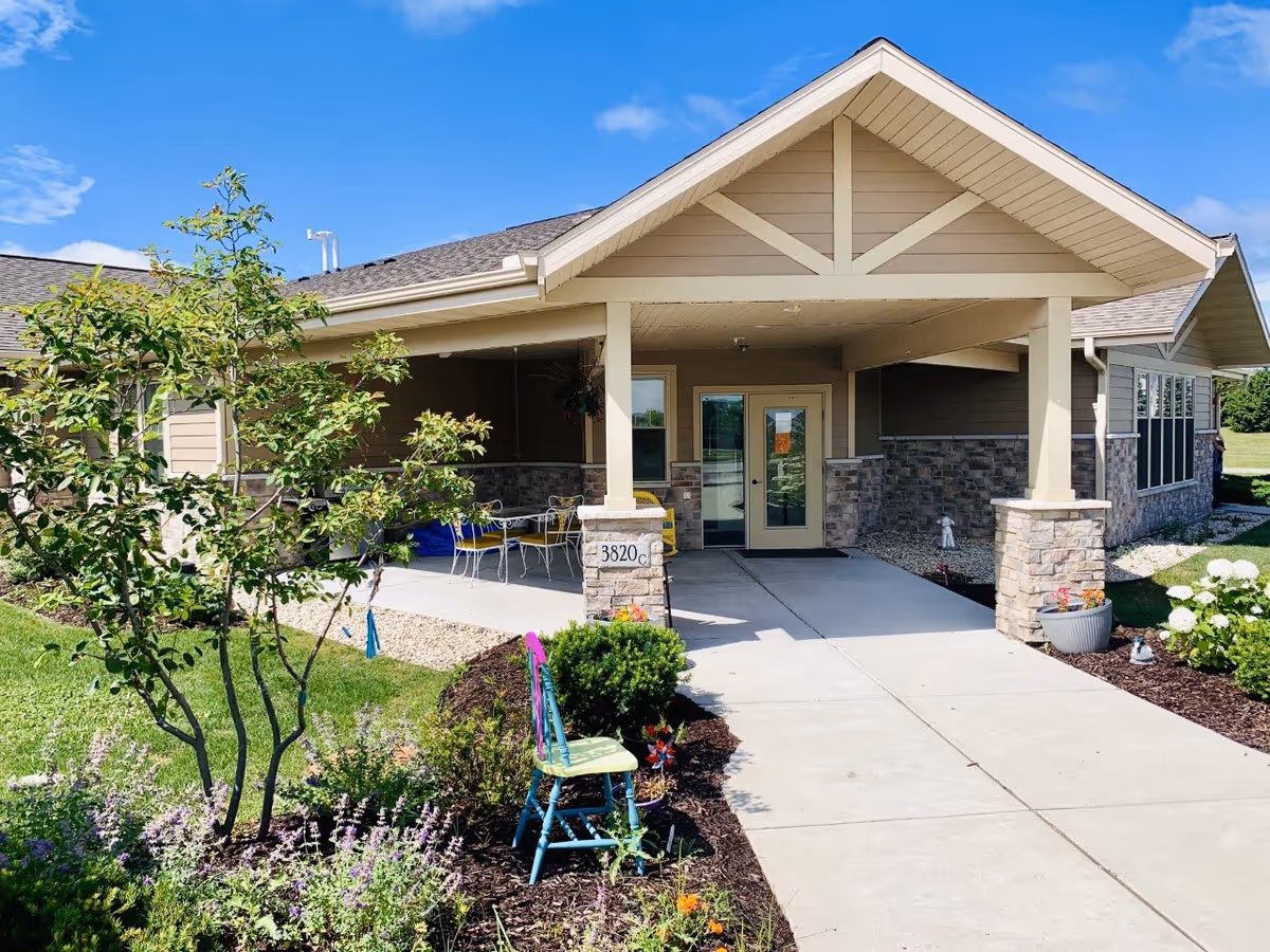 Covered entrance of an assisted living building with a paved walkway, stone pillars, outdoor seating, and landscaped flowerbeds.