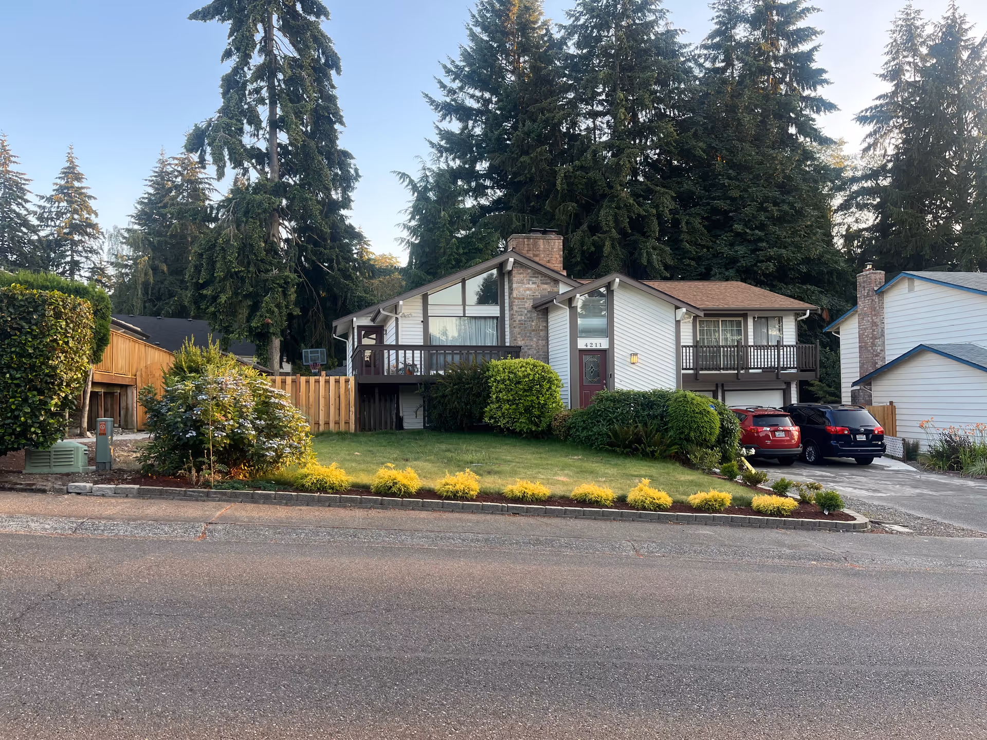 Two-unit house with front lawn, hedges, and a driveway with parked cars.