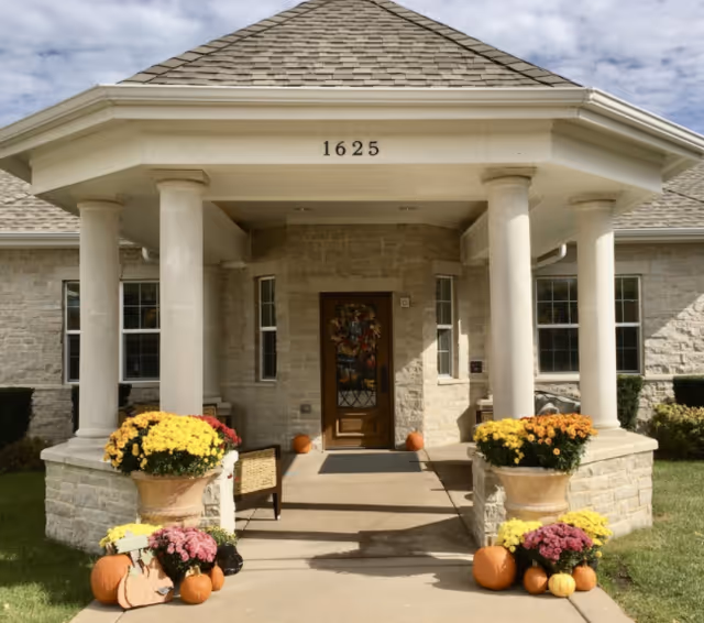 Front entrance of a stone building labeled 1625 with columns, a decorative door, and pumpkins and potted flowers on the walkway.