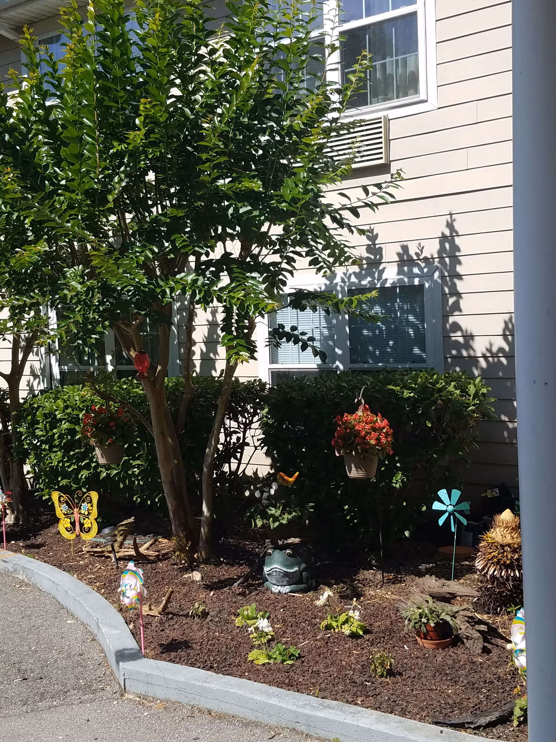 A garden area outside a building with a tree, bushes, and various decorative items including a yellow butterfly ornament, a blue pinwheel, a frog statue, and hanging flower pots with red flowers. The building has beige siding and windows with blinds.