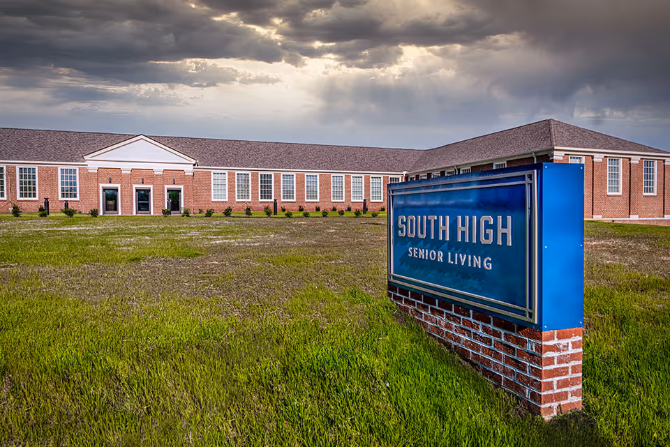 Exterior view of South High Senior Living facility showing a large brick building with many windows under a cloudy sky with sun rays breaking through. In the foreground, there is a blue sign with gold lettering that reads 'South High Senior Living' set on a brick base, surrounded by green grass.