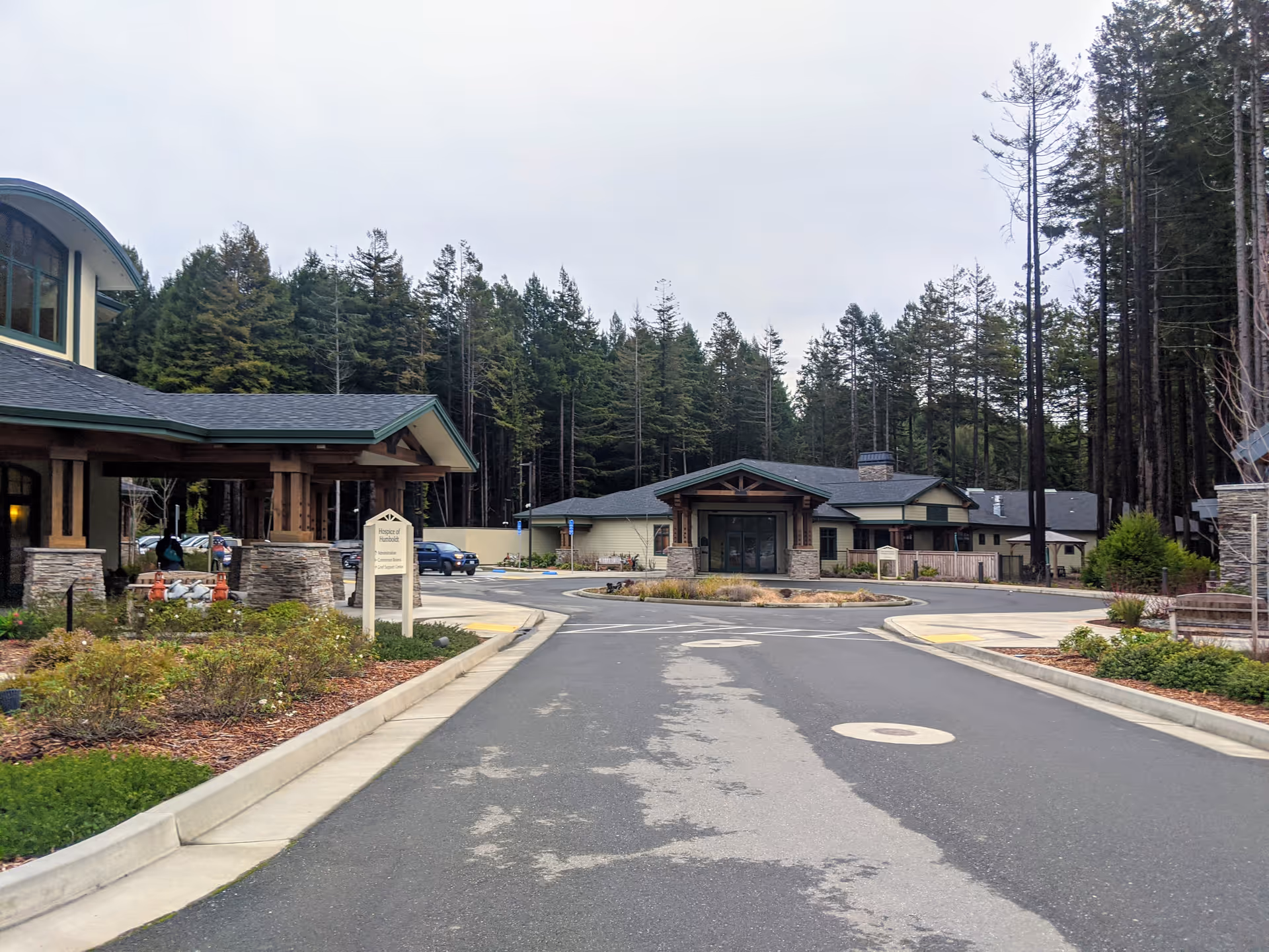 Exterior view of Hospice of Humboldt campus with driveway, entrance canopies, and forest backdrop.