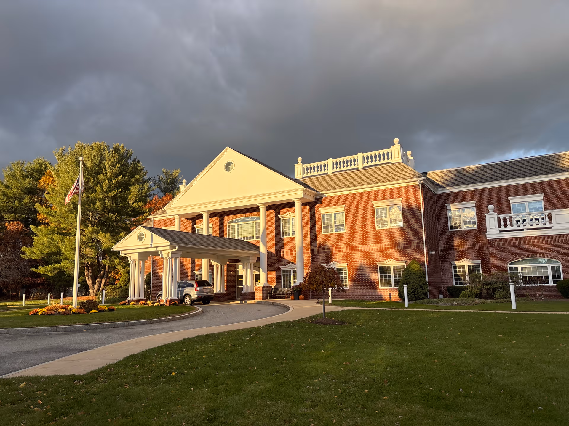 Brick senior living facility with covered entrance, flagpole, and manicured lawn under dark clouds.