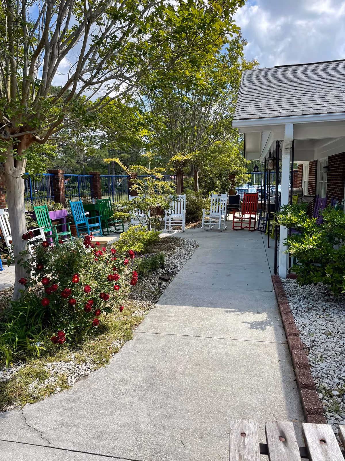 Sunlit outdoor walkway with colorful rocking chairs and landscaped garden beside Elderhaus at the Lake.