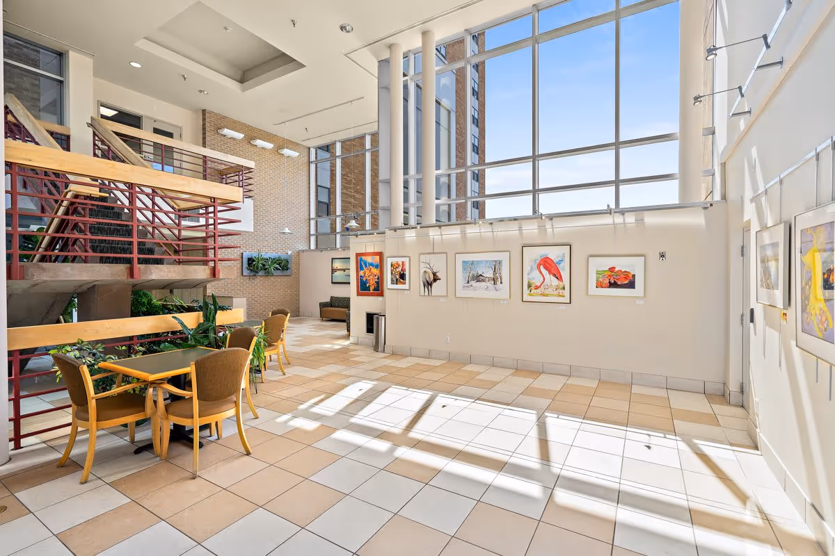 Bright interior common area with large floor-to-ceiling windows letting in natural light. The space features tiled floors, a staircase with red railings, several framed artworks on the walls, and a small seating area with tables and chairs. There are plants placed near the staircase and a brick wall in the background.