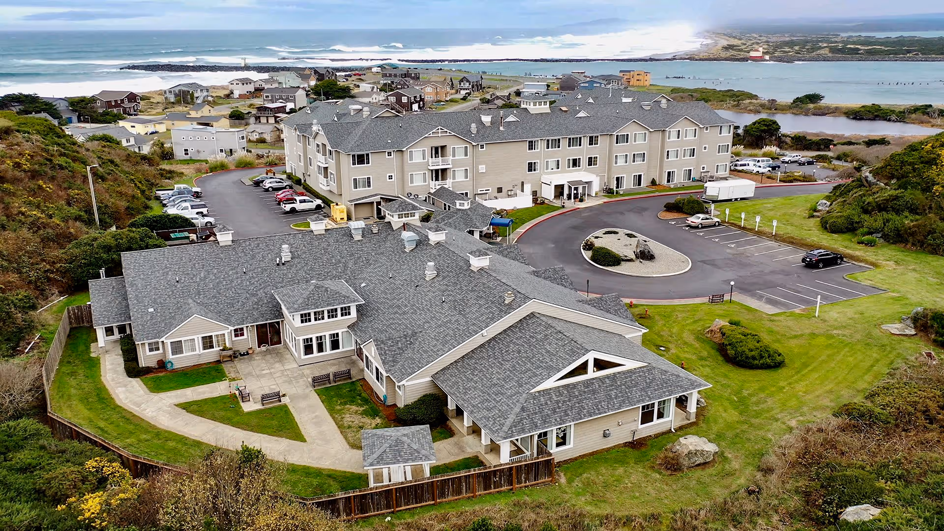 Aerial view of Pacific View Senior Living Community showing two large buildings with gray roofs surrounded by green lawns, a parking lot with several cars, and a scenic ocean coastline in the background.