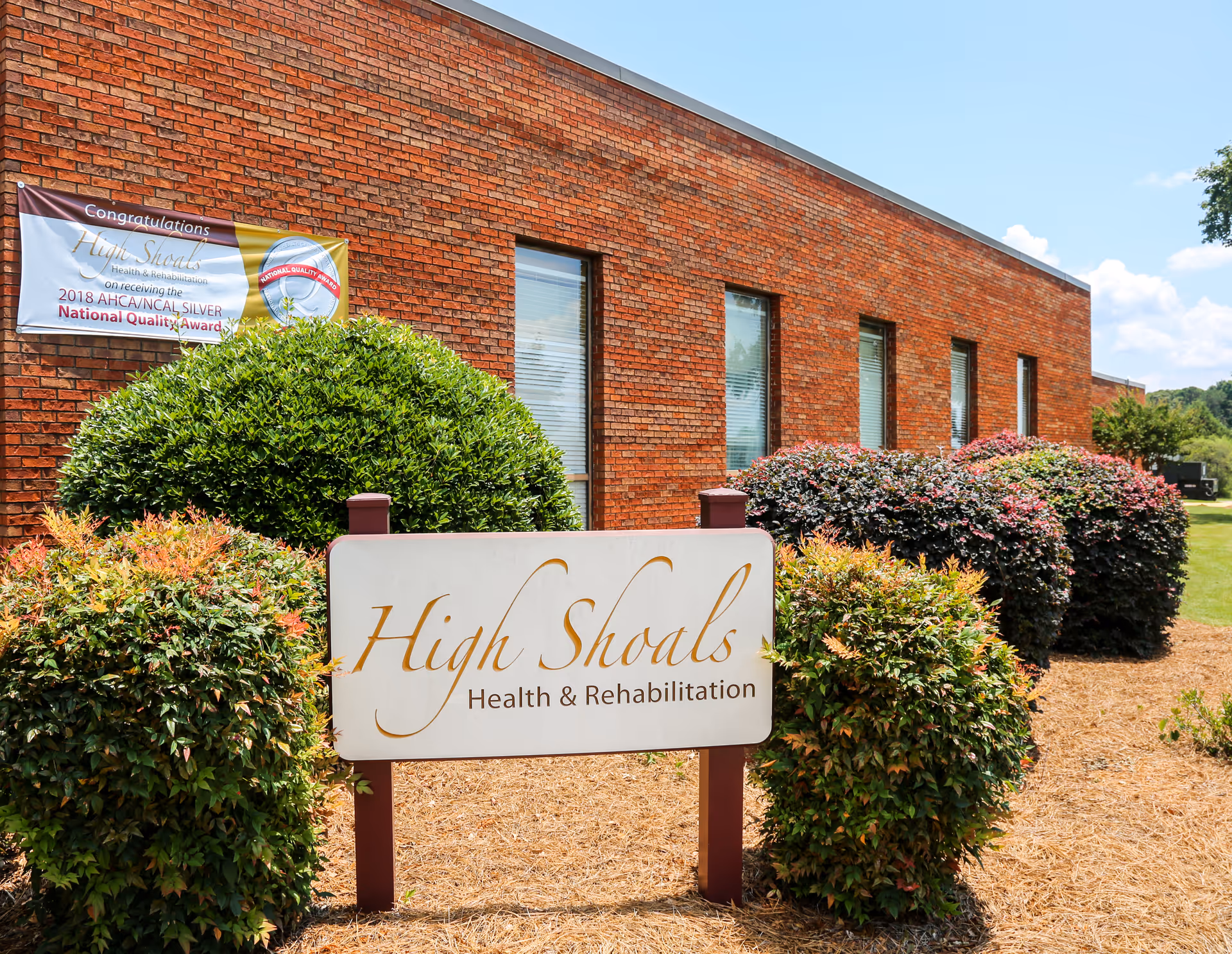 Brick building exterior with a large sign reading High Shoals Health & Rehabilitation, surrounded by shrubs.