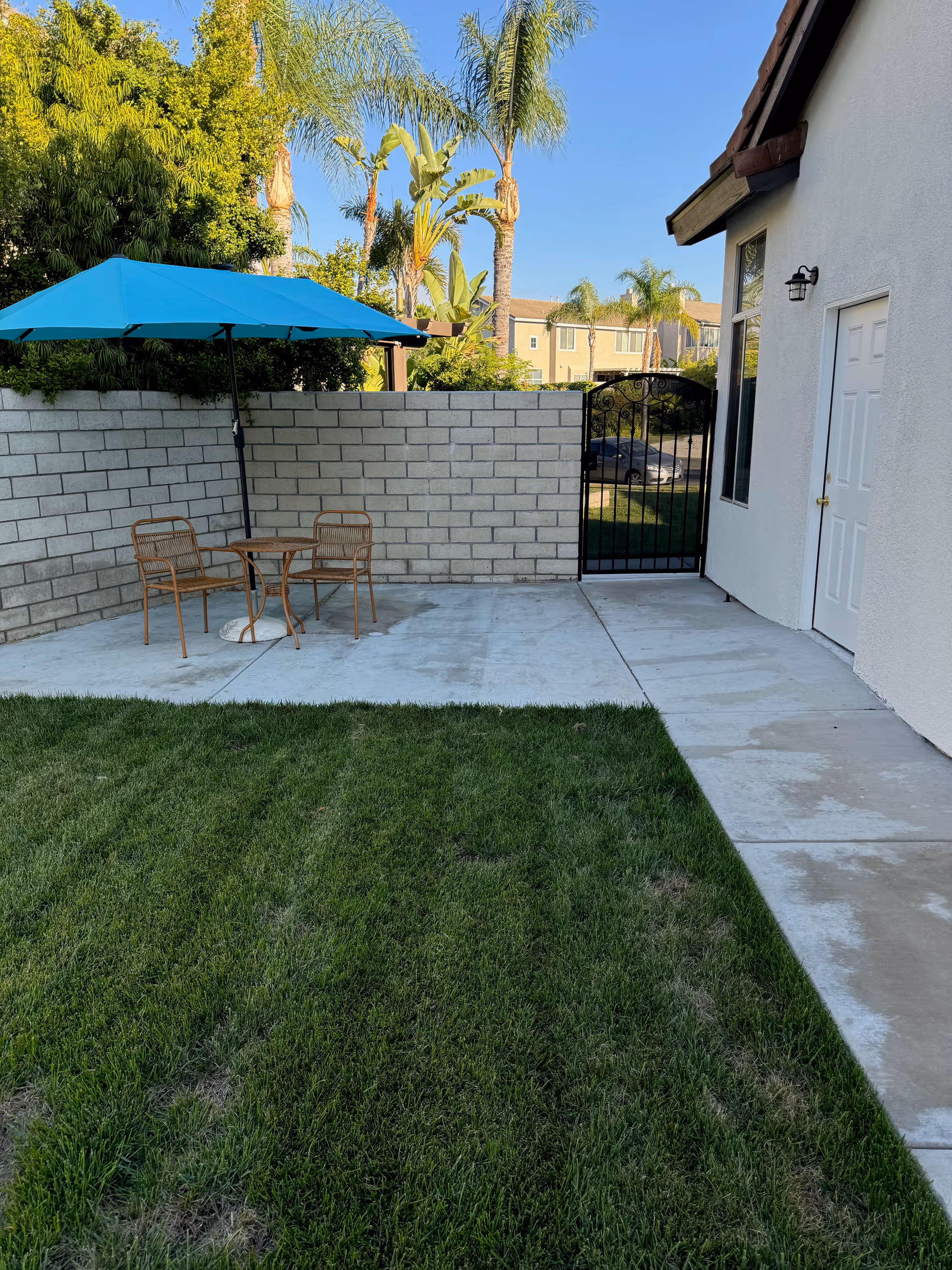 Outdoor courtyard with blue umbrella, table, chairs, and gate beside a white building.