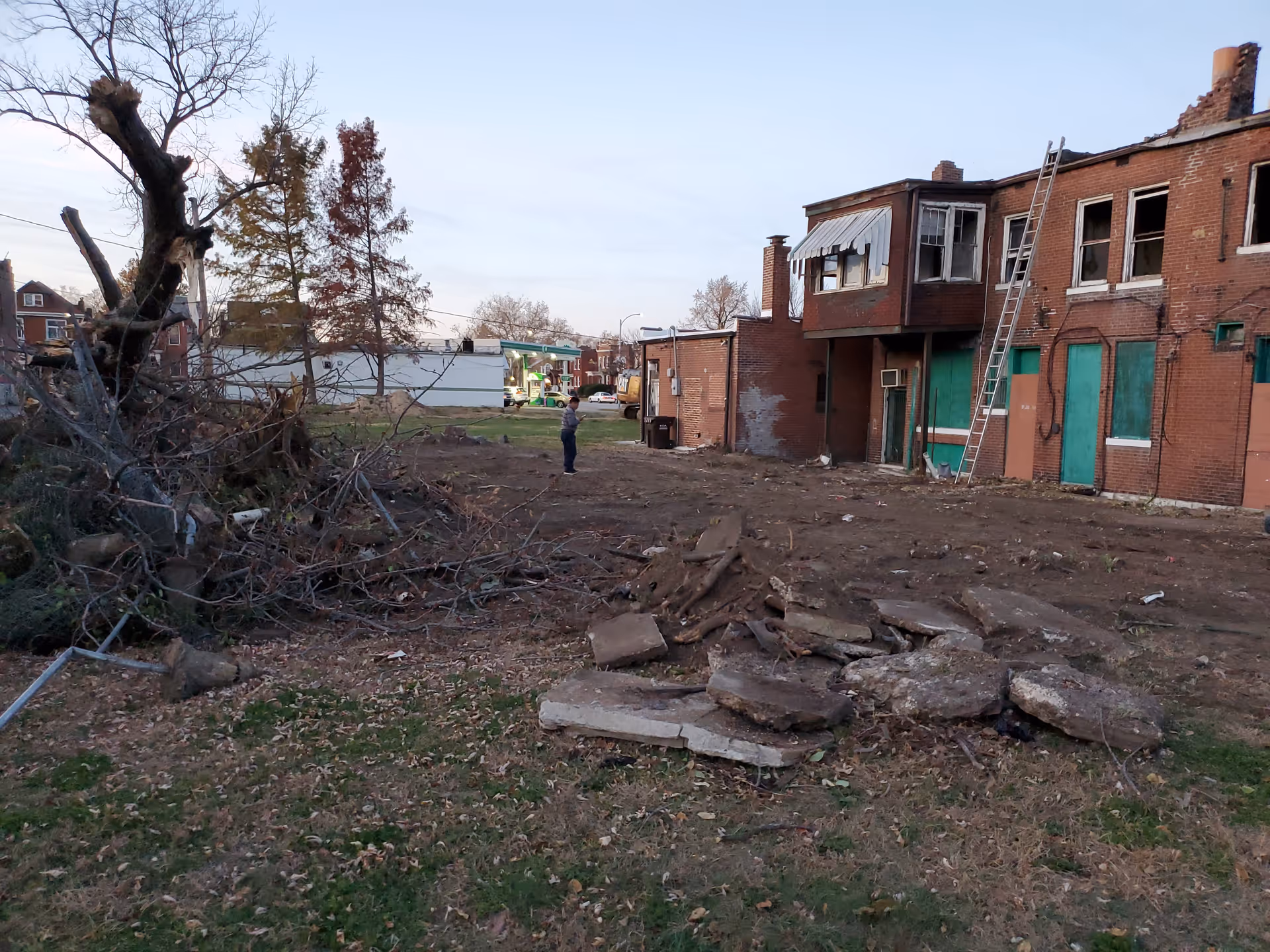 Desolate exterior of a brick building with scattered debris, a ladder, and a man standing nearby.