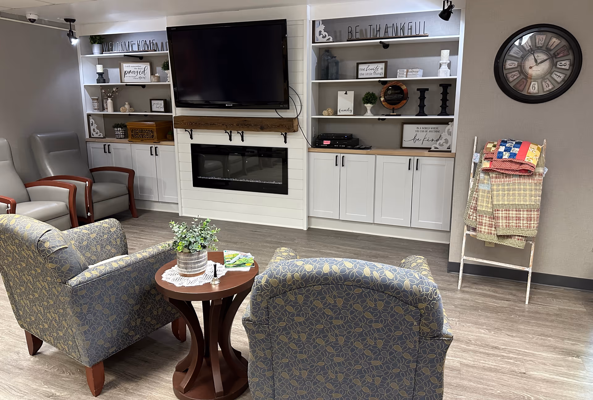 A cozy living room area in a senior care facility with two patterned armchairs and two gray chairs arranged around a small round wooden table with a plant and magazines. The background features built-in white cabinets and shelves decorated with various home decor items, a wall-mounted flat-screen TV above an electric fireplace, a large wall clock, and a wooden quilt rack holding folded quilts.