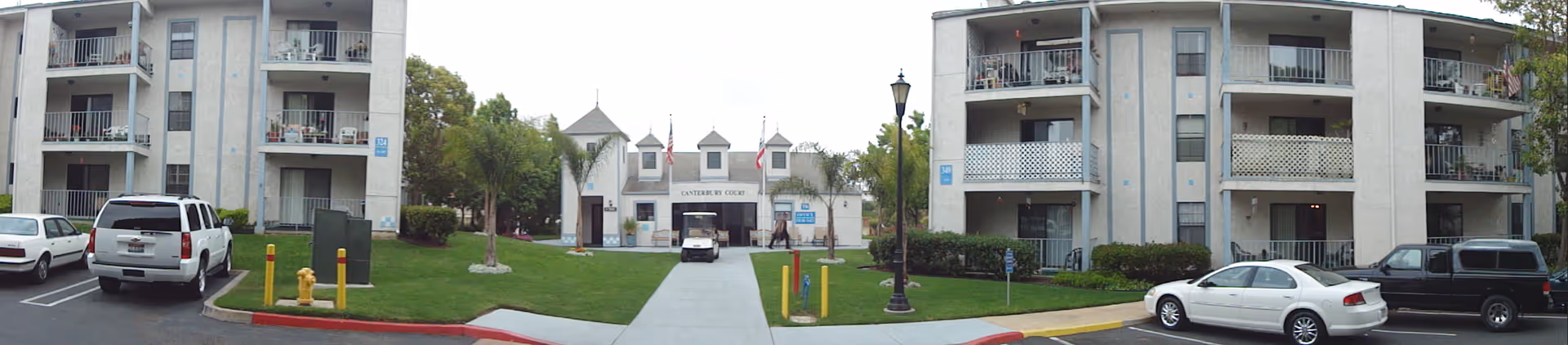 Exterior view of Canterbury Court Senior Apartments showing a three-story residential building on both sides with balconies. In the center is a pathway leading to the main entrance of the facility, which has a white building with a peaked roof and three small towers. There are several parked cars and a golf cart near the entrance, with green lawns and palm trees lining the walkway.
