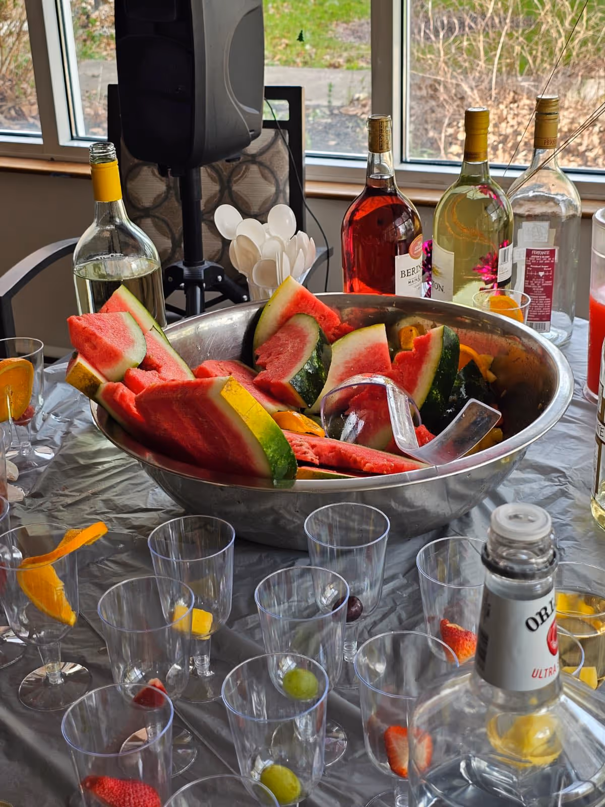 Large bowl of sliced watermelon and other fruit surrounded by plastic cups and bottles on a table by a window.